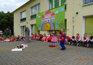 Taras przedszkolny, a na nim scenografia przedstawiająca zamek. Na scenie dwoje dzieci stojących oraz jedno leżące na kocyku.
